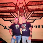 two muscular gay bear men wearing QRLY Merch graphic t-shirts posing together in front of a decorative wall at a bar