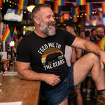 gay bear man wearing a black ‘feed me to the bears’ t-shirt sitting at a bar decorated with rainbow pride flags