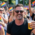 Smiling man holding a beer at an outdoor Pride parade with rainbow flags in the background.
