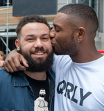two gay men smiling as one kisses the other on the cheek, wearing a white QRLY Merch shirt outdoors