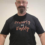 smiling bearded man wearing a black ‘Property of Daddy’ QRLY Merch t-shirt indoors.