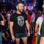 muscular bearded man wearing a black QRLY Merch leather-style graphic t-shirt at a bar with rainbow lights