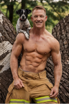 sexy firefighter calendar photo featuring shirtless muscular man with dog in outdoor forest setting for gay men’s calendar